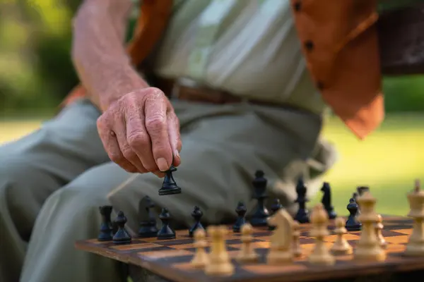 Close up of senior man playing chess in a city park, holding chess piece in hand.
