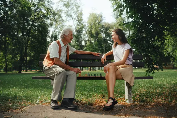 A young caregiver spending quality time with lonely senior client in the city park. The elderly man spending time outdoors with his granddaughter, playing chess and chatting.