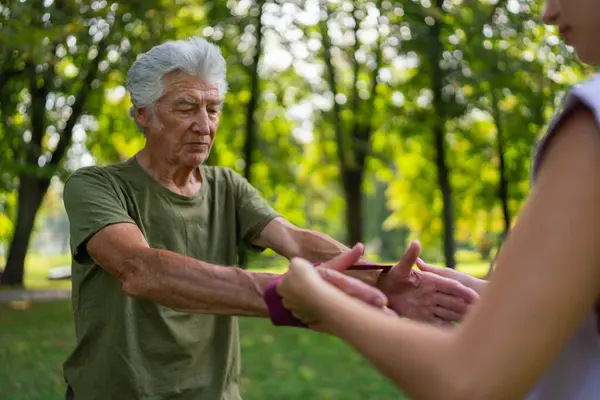 The senior man exercising in the park, using resistance band. A young caregiver showing older man how to perform the exercise with resistance band correctly. A portrait of a young physiotherapist