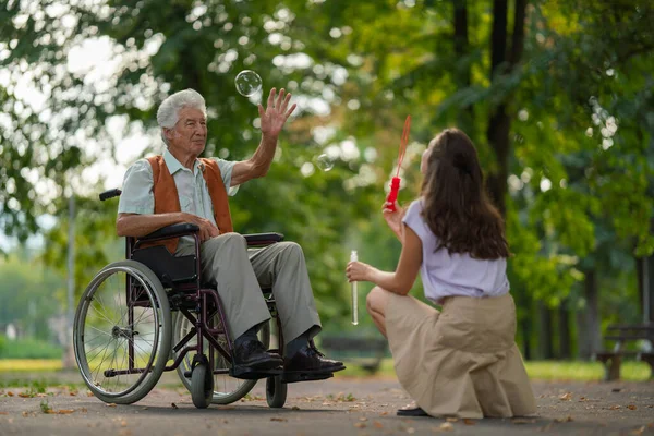 車椅子の男が公園の介護者と楽しんでバブルを捕まえました 車椅子のシニアマンは 若い看護師によって吹き飛ばされた泡をキャッチするように動き 簡単な演習を行います — ストック写真