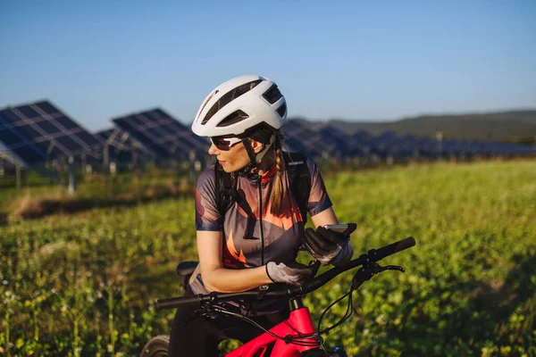 Portrait of a beautiful cyclist standing in front of solar panels at a solar farm during a summer bike tour in nature. A solar farm as solution for more sustainable energy future.