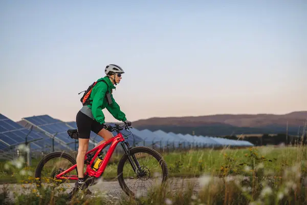 自然の中で夏の自転車ツアー中にソーラーファームでソーラーパネルの前に乗る美しいサイクリストの側面 より持続可能なエネルギーの未来のためのソリューションとしてのソーラーファーム — ストック写真