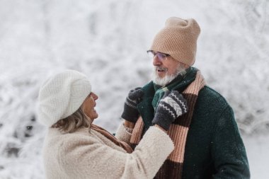 Elegant senior couple walking in the snowy park, during cold winter snowy day. Elderly woman fasten husbands winter coat. Wintry landscape. clipart