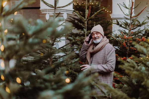 Senior man standing among Christmas trees, deciding which one to buy. Elderly man making call with smart phone.