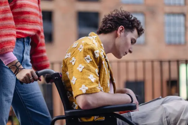 Sad gen Z boy in a wheelchair with friends in the city. Inclusion, equality, and diversity among Generation Z. Sadness and helplessness in the face of chronic illness.