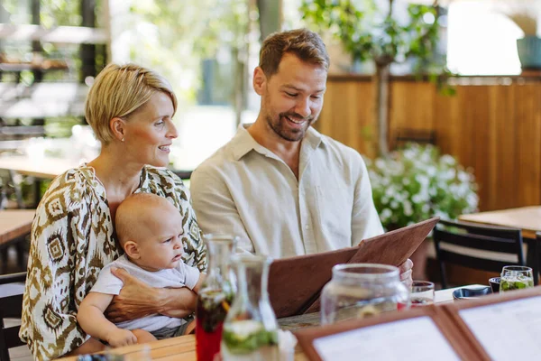 Family with baby reading menu in a restaurant, choosing food and drinks. Mother holding baby on knees during lunch. Family dinner at a restaurant.