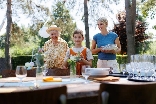 おばあちゃん お母さんと娘が夏の庭のパーティーのテーブルを用意しています プレート 食べ物 飲み物を持参する — ストック写真