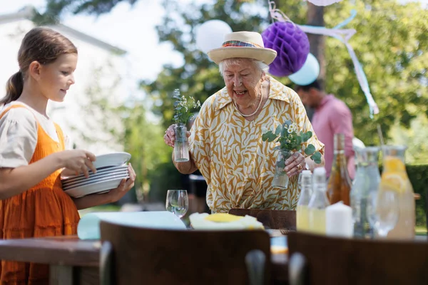 サマーガーデンパーティーのための祖母と孫の設定テーブル プレートと花の装飾を持って来る — ストック写真