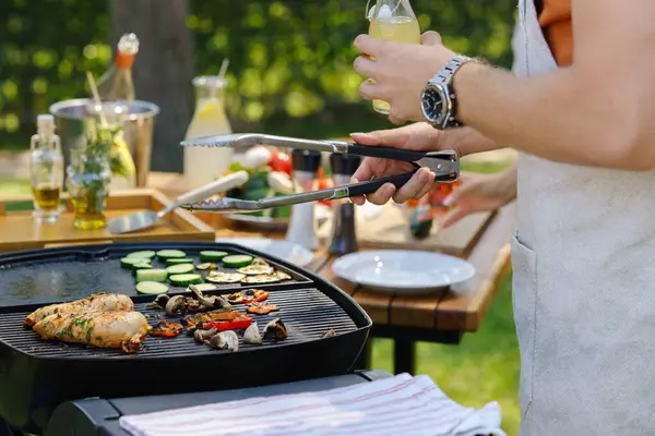 Close up of a man grilling at a bbq garden party. Grill master holding barbecue tongs and glass with fresh lemonade.