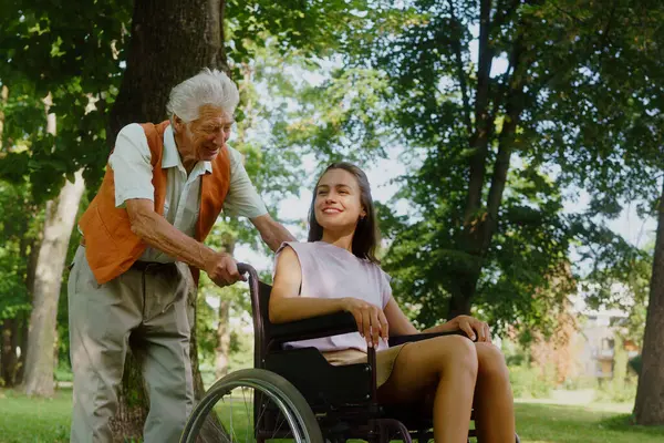 The senior man pushing young caregiver in a wheelchair, having fun. The elderly man feeling good and fit, he is able to push his granddaughter in the wheelchair.