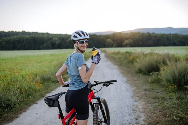 Diabetic cyclist with a continuous glucose monitor on her arm drinking water during her bike tour to manage her diabetes while exercising. Concept of exercise and diabetes.