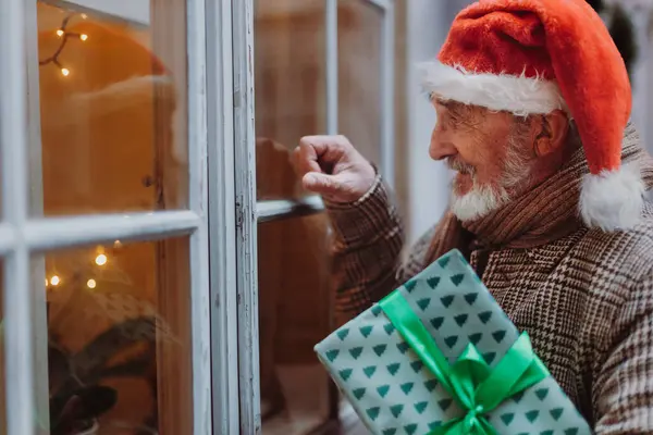 Senior man with a Christmas gift in hand standing in front of the window and knocking on it. The elderly man with christmas hat playing Santa Claus, bringing a gift.