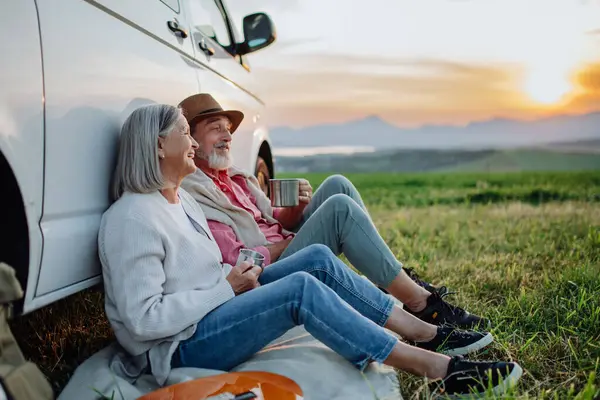 Potrait of senior couple sitting by car and drinking coffee after long drive during their roadtrip. Elderly spouses at autumn roadtrip watching the sun set behind High Tatras.