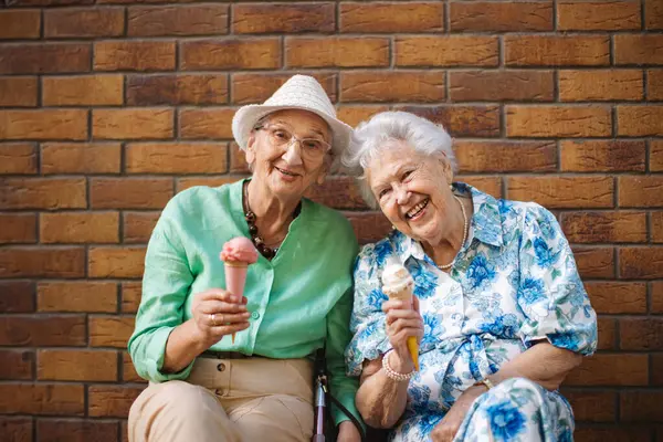 Portrait of two senior female friends in the city, eating ice cream on a hot summer day. Elderly ladies on summer vacation in the city. Pensioners on group trip. Concept of senior vacation and travel