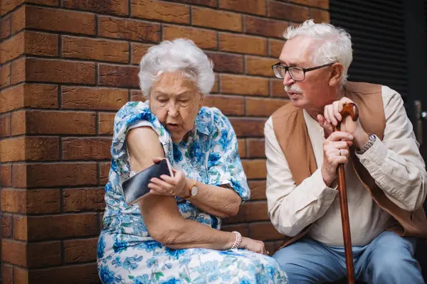 Diabetic senior woman checking blood glucose level with continuous glucose monitor outdoors in the city. Elderly husband waiting while his chronically ill wife connecting her CGM with smarphone.