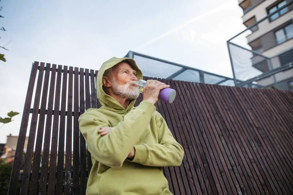 Senior man exercising outdoors in the city. Elderly man drinking water after his morning run. Low angle shot.