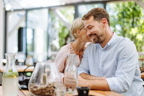 Portrait of beautiful couple in a restaurant, on a romantic date. Wife whispers into his husbands ear, having a romantic moment at restaurant patio.