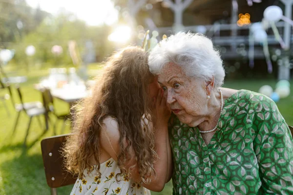 在一个花园的宴会上 小女孩在祖母耳边低声说着一个秘密 祖父母与孙子孙女之间的爱与亲密 — 图库照片