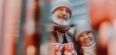 Happy senior couple enjoying outdoor christmas market at the evening, drinking muled wine from christmas mugs. Christmas banner with copy space. clipart