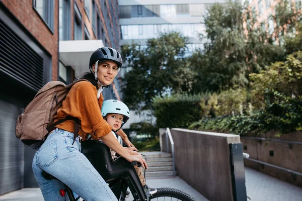 Mother carring her son on a secure child bike carrier or seat, both wearing helmets. Mom commuting with a young child through the city on a bicycle.