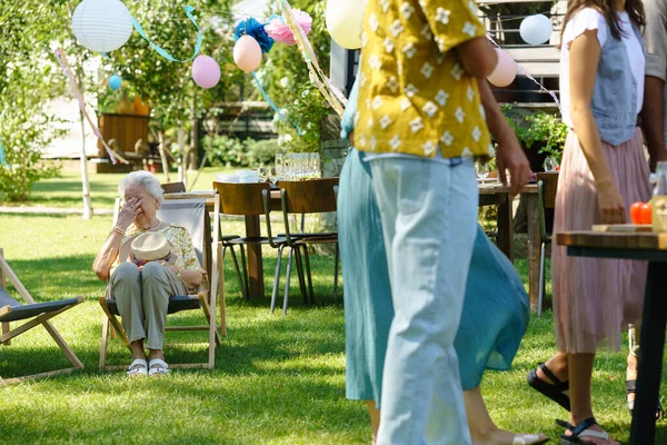 Tired elderly woman sitting on a folding chair at a garden party, taking a break after a long outdoor grill party. Elderly woman cries tears of joy, happy to be with her family and loved ones.