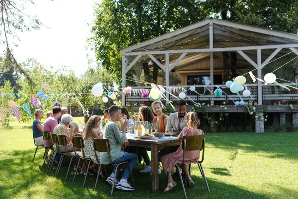 Family and friends sitting at the party table during a summer garden party outdoors. Decorations and the beautiful wooden gazebo at background.