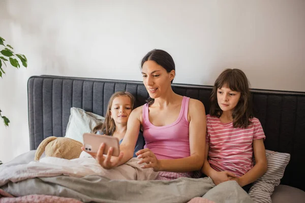 毛布の下でベッドに横たわっている2人の娘と母親の肖像画 家族がお父さんにスマートフォンでビデオ通話をする 母の日と母の愛の概念 — ストック写真