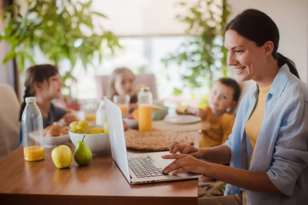 Mother working on a laptop at the kitchen table while the children have breakfast. Remote work and home office for mothers with children.