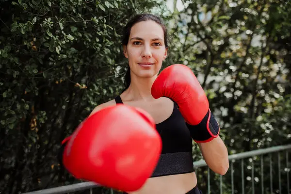 Portrait of confident boxer woman in red boxing gloves standing outdoors. Woman boxing for relaxation and bolsting mental health. Boxing empowers women.