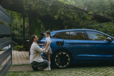 Aile arabalarını evin önünde şarj ederken baba-kız konuşmalarının portresi. Şarj limanında şarj aleti olan elektrikli bir araç. Baba kızını okula götürüyor..