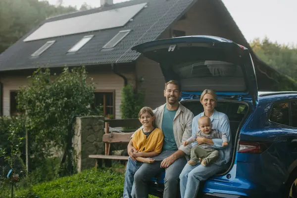 Family sitting in the trunk of electric car, with house with solar panels on roof behind them. Solar energy and sustainable lifestyle of young family. Concept of green energy and sustainable future