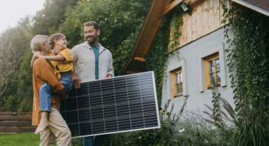 Aile bahçede güneş paneli tutarak yürüyor. Güneş enerjisi ve genç ailenin sürdürülebilir yaşam tarzı. Yeşil enerji kavramı ve gelecek nesiller için sürdürülebilir gelecek.