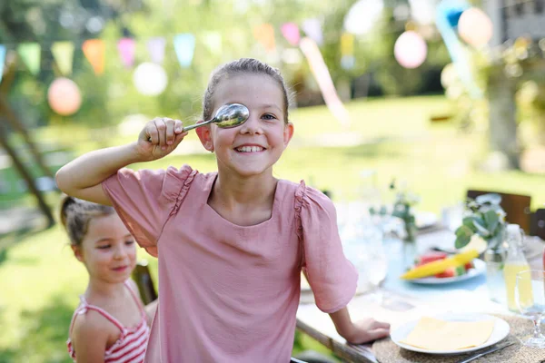 Girl covering eye with a spoon in front of her face. Friends are having fun at childrens birthday party. Cousins having fun at a family garden party outdoors.