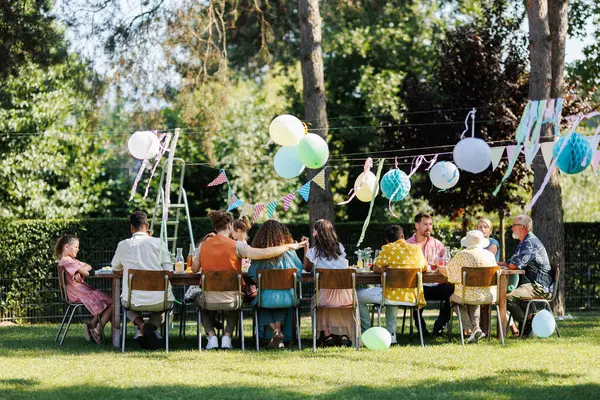Family and friends sitting at the party table during a summer garden party outdoors. Rear view of people sitting at the table outdoors.