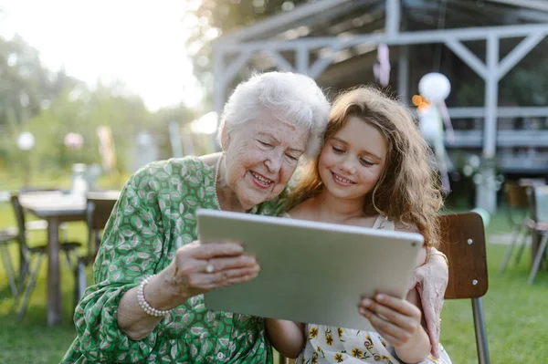 Young girl showing someting on tablet to elderly grandmother at a garden party. Love and closeness between grandparent and grandchild.