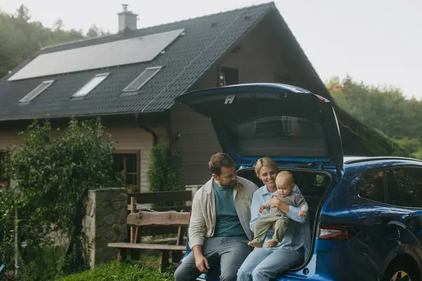 Family sitting in the trunk of electric car, with house with solar panels on roof behind them. Solar energy and sustainable lifestyle of young family. Concept of green energy and sustainable future