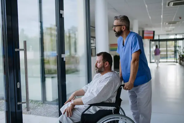 病院の廊下に車椅子で患者を押している男性看護師 過体重の患者は不安を感じ 健康上の懸念を持っています — ストック写真