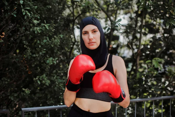 Portrait of muslim boxer woman in red boxing gloves and hijab standing outdoors. Woman boxing for relaxation and bolsting mental health. Boxing empowers women.