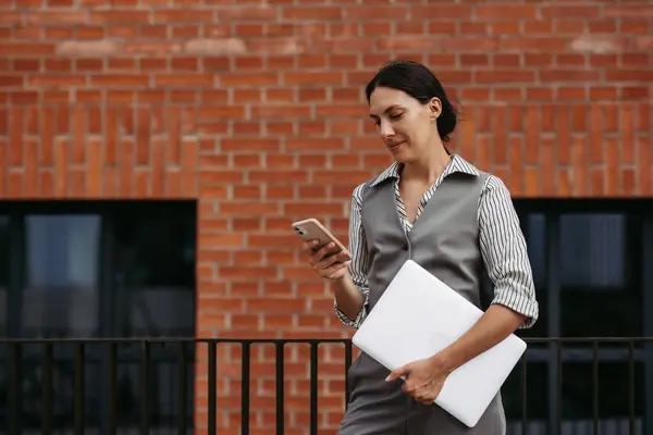 Portrait of beautiful woman in business attire standing on city street. Muslim businesswoman with laptop making call. Female university professor in front of university building.