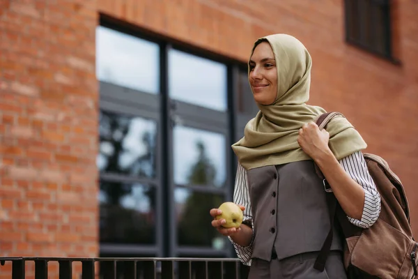 Portrait of beautiful woman in hijab standing on city street. Muslim studnet eating apple, going to school. Iran, Afganistan female teacher in front of school building.