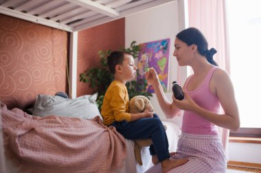 Genç, güzel bir anne ilaç veriyor, kaşıktan kaşığa öksürük şurubu döküyor hasta oğluna. Çocukların mevsimsel hastalıklar, virüsler ve çocukların bağışıklık sistemi kavramı.