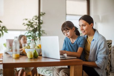 Anne, çocuklar kahvaltı ederken mutfak masasında dizüstü bilgisayarla çalışıyor. Çocuklu anneler için uzak iş ve ev ofisi.