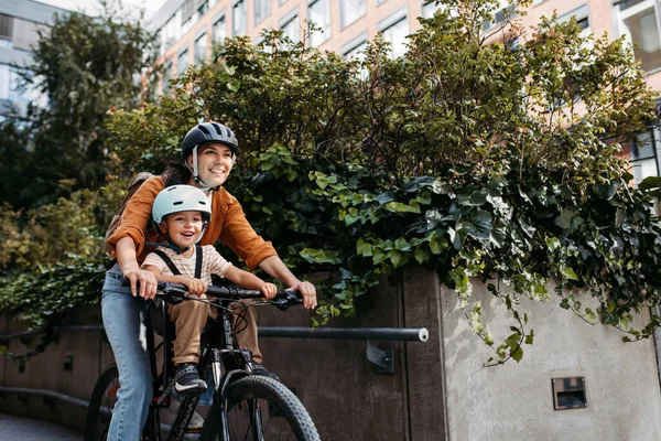母亲带着她的儿子坐在安全的儿童自行车载体上或座位上 两个人都戴着钢盔 妈妈和一个小孩骑自行车在城市里穿梭 — 图库照片