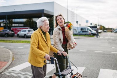 Mature granddaughter carrying grandmothers shopping bag. Senior woman and caregiver going to home with goceries from the supermarket, during cold autumn day. clipart