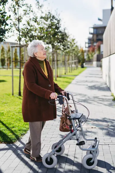 A senior woman with a mobility walker walking on the city streets during autumn day, enjoying the beautiful sunny weather. Elderly lady savoring every moment, living life to fullest.