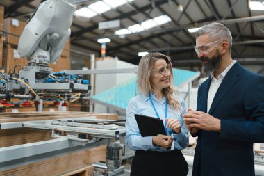 Kadın mühendis ve erkek proje yöneticisi modern endüstriyel fabrikada duruyor, ahşap mobilya üretiminden bahsediyorlar. Robotik, robotik kolları olan büyük mobilya üretim tesisi.