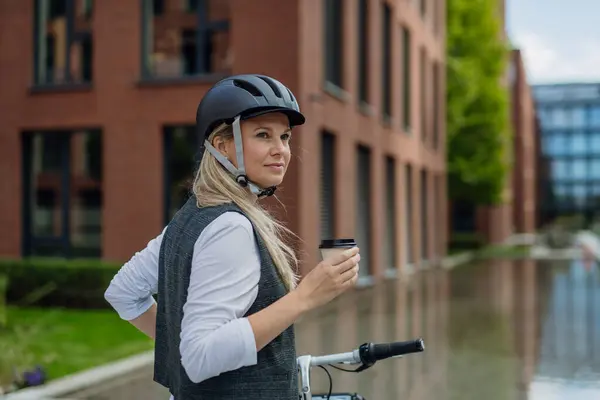Beautiful middle-aged woman commuting through the city, buying, drinking coffe in front of office. Female city commuter with helmet traveling from work by bike after a long workday.