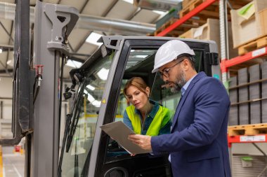 Kadın forklift sürücüsü takım elbiseli depo müdürüyle konuşuyor, sipariş seçiyor. Depo işçisi nakliye, teslimat ve depodaki stokları kontrol etmek için ürünler hazırlıyor..