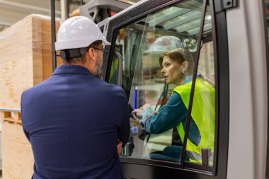 Kadın forklift sürücüsü takım elbiseli bir depo müdürüyle konuşuyor. Depo işçisi nakliye, teslimat ve depodaki stokları kontrol etmek için ürünler hazırlıyor..