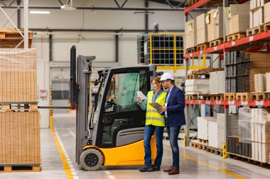 Kadın forklift şoförü, depo çalışanı müdürüyle konuşuyor, forkliftin yanında bekliyor. Depo işçisi nakliye, teslimat ve depodaki stokları kontrol etmek için ürünler hazırlıyor..